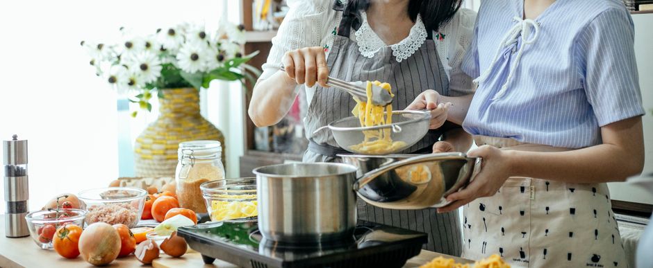 Unrecognizable female cooks putting pasta into sieve with tongs while standing near table with pan on stove and various ingredients while cooking lunch
